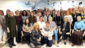 Eine Gruppe von etwa 50 Personen steht und sitzt in einem Raum, umgeben von einer Wand mit Vogelmotiven.