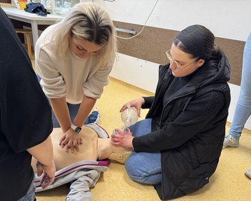 Zwei Frauen führen eine Herz-Lungen-Wiederbelebung an einer Übungspuppe durch, während eine weitere Person zuschaut.