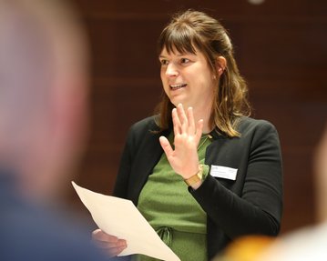 Eine Frau mit langen, braunen Haaren hält ein Blatt Papier in der Hand und spricht, während sie in einem formellen Umfeld steht.