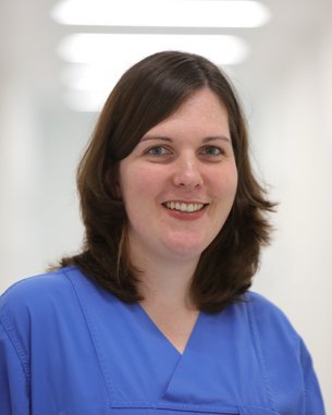 Eine Frau mit braunen Haaren trägt eine blaue medizinische Uniform. Sie lächelt freundlich und steht in einem hellen, modernen Flur. Der Hintergrund ist unscharf, was den Fokus auf die Frau lenkt.