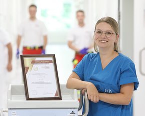 Eine Frau in blauer Arbeitskleidung steht neben einem Rollwagen mit einem Zertifikat. Im Hintergrund sind zwei Personen in Arbeitskleidung sichtbar.