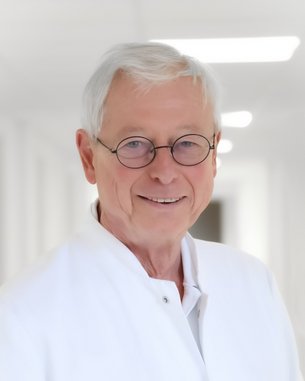 Ein älterer Arzt mit Brille und weißem Kittel lächelt freundlich in einem hellen Flur eines Krankenhauses.