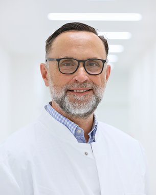 Ein lächelnder Arzt mit Brille und Bart trägt einen weißen Kittel und steht in einem hellen Flur eines medizinischen Umfelds.