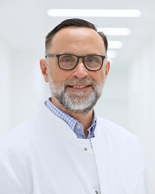 Ein lächelnder Arzt mit Brille und Bart trägt einen weißen Kittel und steht in einem hellen Flur eines medizinischen Umfelds.