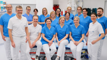 Eine Gruppe von 17 Personen in blauen und weißen Arbeitskleidung, die in einem medizinischen Raum posieren.