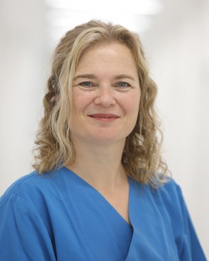 Eine Frau mit lockigem, blonden Haar trägt eine blaue medizinische Uniform. Sie lächelt freundlich in die Kamera. Der Hintergrund ist unscharf und hell, was eine klinische Umgebung suggeriert.