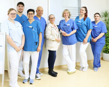 Eine Gruppe von acht medizinischen Fachkräften in blauen und weißen Uniformen steht lächelnd vor einem Schild, das zur Inneren Medizin Einheit weist.