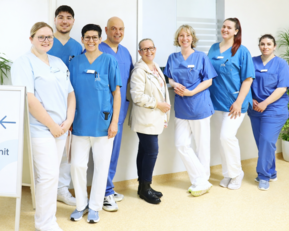 Eine Gruppe von acht medizinischen Fachkräften in blauen und weißen Uniformen steht lächelnd vor einem Schild, das zur Inneren Medizin Einheit weist.