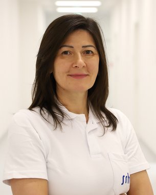 Eine Frau mit langen, dunklen Haaren trägt ein weißes Poloshirt und steht in einem hellen Flur mit neutralem Hintergrund.