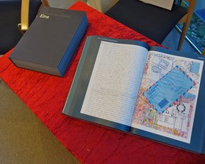 Ein aufgeschlagenes Buch mit handschriftlichem Text und einer farbigen Karte liegt auf einem roten Tisch. Daneben befindet sich eine graue Box mit dem Titel "Eins".