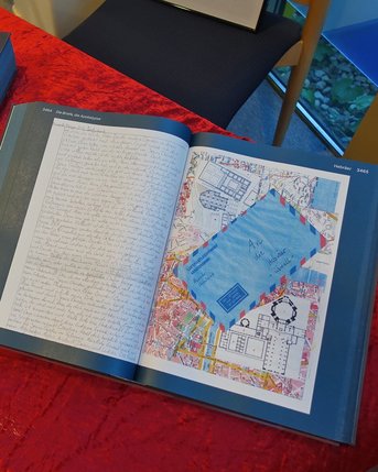 Ausstellung eines Druckexemplars der St. Galler Corona-Bibel. Ein aufgeschlagenes Buch mit handschriftlichem Text und einer farbigen Karte liegt auf einem roten Tisch. Daneben befindet sich eine graue Box mit dem Titel "Eins".