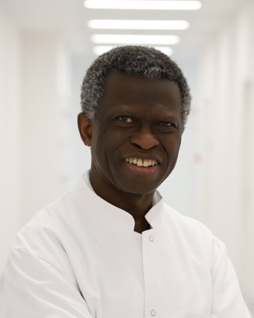 Ein lächelnder Arzt mit grauen Haaren trägt einen weißen Kittel und steht mit verschränkten Armen in einem hellen Krankenhausflur.