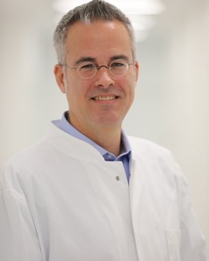 Ein Mann in einem weißen Kittel mit Brille lächelt freundlich in einem hellen, modernen Raum.