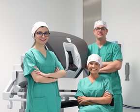 Drei medizinische Fachkräfte in grüner OP-Kleidung stehen und sitzen vor einem chirurgischen Roboter in einem modernen Operationssaal.