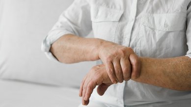 Eine Person sitzt und hält ihren Handgelenk, möglicherweise aufgrund von Schmerzen oder Unbehagen. Sie trägt ein weißes Hemd.