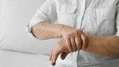 Eine Person sitzt und hält ihren Handgelenk, möglicherweise aufgrund von Schmerzen oder Unbehagen. Sie trägt ein weißes Hemd.