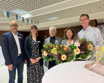 Fünf Personen stehen zusammen und halten einen großen Blumenstrauß. Sie tragen formelle Kleidung und lächeln in die Kamera.