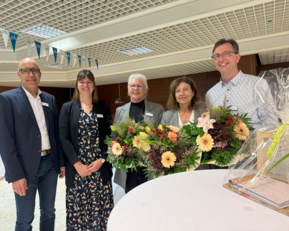 Fünf Personen stehen zusammen und halten einen großen Blumenstrauß. Sie tragen formelle Kleidung und lächeln in die Kamera.