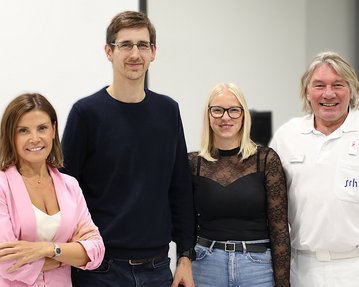 Vier Personen stehen zusammen in einem modernen Raum. Sie lächeln und tragen unterschiedliche Kleidung.