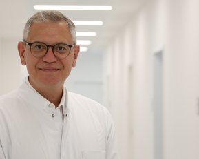 Ein Arzt in einem weißen Kittel steht in einem hellen Flur mit mehreren Türen im Hintergrund.