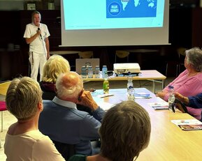 Ein Referent präsentiert Informationen über "Blue Zones" vor einer Gruppe älterer Menschen in einem Seminarraum.