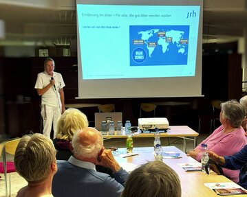Ein Referent präsentiert Informationen über "Blue Zones" vor einer Gruppe älterer Menschen in einem Seminarraum.