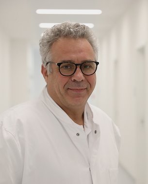 Ein Arzt in einem weißen Kittel steht in einem hellen Flur. Er trägt eine Brille und lächelt leicht.