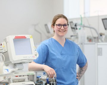 Eine Krankenschwester in blauer Arbeitskleidung steht lächelnd neben medizinischen Geräten in einem Krankenhausumfeld.