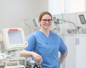 Eine Krankenschwester in blauer Arbeitskleidung steht lächelnd neben medizinischen Geräten in einem Krankenhausumfeld.