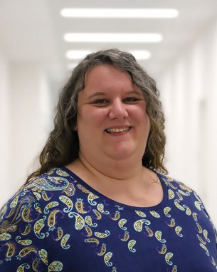 Eine Frau mit langen, lockigen Haaren trägt ein blaues Oberteil mit einem bunten Muster. Sie lächelt und steht in einem hellen Flur.