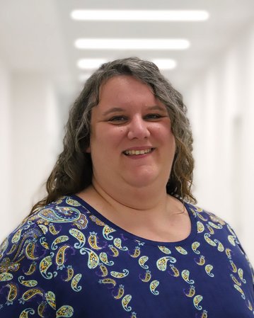 Eine Frau mit langen, lockigen Haaren trägt ein blaues Oberteil mit einem bunten Muster. Sie lächelt und steht in einem hellen Flur.