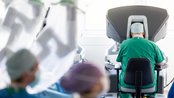 Ein Chirurg in grüner Kleidung sitzt an einem Operationsroboter, umgeben von weiteren medizinischen Fachkräften in OP-Kleidung.
