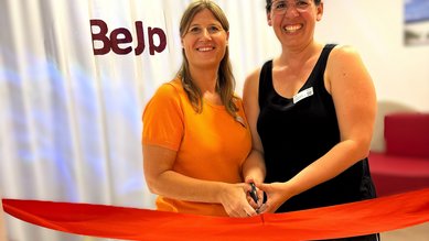 Zwei Frauen halten eine Schere und stehen vor einem roten Band, das sie durchschneiden. Im Hintergrund ist ein Banner mit der Aufschrift "BeJp".
