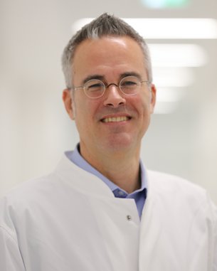 Ein lächelnder Arzt in einem weißen Kittel steht in einem hellen Flur. Er trägt eine Brille und hat graue Haare.