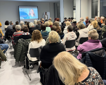 Eine große Gruppe von Menschen sitzt in einem Raum und hört einem Vortrag zu, während eine Präsentation auf einer Leinwand gezeigt wird.