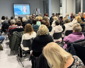 Eine große Gruppe von Menschen sitzt in einem Raum und hört einem Vortrag zu, während eine Präsentation auf einer Leinwand gezeigt wird.