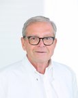Ein älterer Mann mit Brille trägt einen weißen Kittel. Er steht vor einem hellen Hintergrund und schaut direkt in die Kamera.