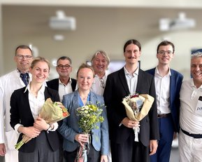Eine Gruppe von neun Personen, die Blumensträuße halten, posiert zusammen in einem hellen Raum. Einige tragen formelle Kleidung, andere laborkittel.