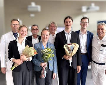 Eine Gruppe von neun Personen, die Blumensträuße halten, posiert zusammen in einem hellen Raum. Einige tragen formelle Kleidung, andere laborkittel.
