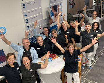 Eine Gruppe von Mitarbeitern steht in einem Empfangsbereich, lächelt und hebt die Hände. Sie tragen einheitliche schwarze Shirts und weiße Hosen.