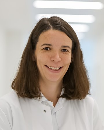 Eine Frau mit langen, braunen Haaren trägt einen weißen Kittel und lächelt freundlich. Der Hintergrund ist hell und unscharf, was den Fokus auf die Person lenkt.
