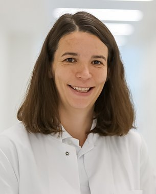 Eine Frau mit langen, braunen Haaren trägt einen weißen Kittel und lächelt freundlich. Der Hintergrund ist hell und unscharf, was den Fokus auf die Person lenkt.