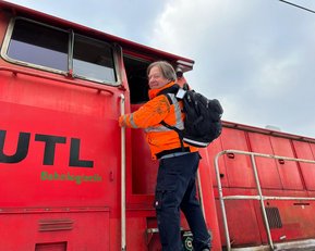 Ein Mann in orangefarbener Arbeitskleidung steigt in eine rote Lokomotive ein, während er einen Rucksack trägt.