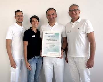 Vier Personen in weißen Outfits halten ein Zertifikat mit der Aufschrift "ZERTIFIKAT". Sie stehen vor einer neutralen Wand.