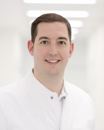 Ein lächelnder Mann in einem weißen Kittel steht in einem hellen, modernen Raum.