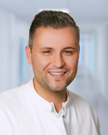 Ein lächelnder Mann in einem weißen Kittel, der in einem hellen, modernen Krankenhausflur steht.