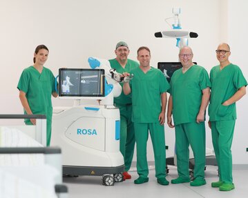 Fünf medizinische Fachkräfte in grüner Kleidung stehen um einen chirurgischen Roboter namens ROSA in einem modernen Operationssaal.