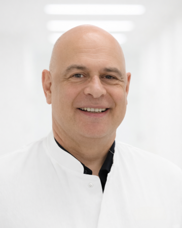 Ein lächelnder Arzt in einem weißen Kittel steht in einem hellen, modernen Krankenhausflur.