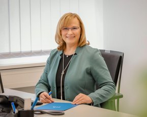 Eine Frau mit blonden Haaren sitzt an einem Schreibtisch, trägt eine grüne Jacke und lächelt, während sie einen Stift in der Hand hält.