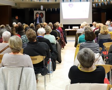 Eine große Gruppe älterer Menschen sitzt in einem Raum und schaut auf eine Präsentation, die an einer Leinwand gezeigt wird.